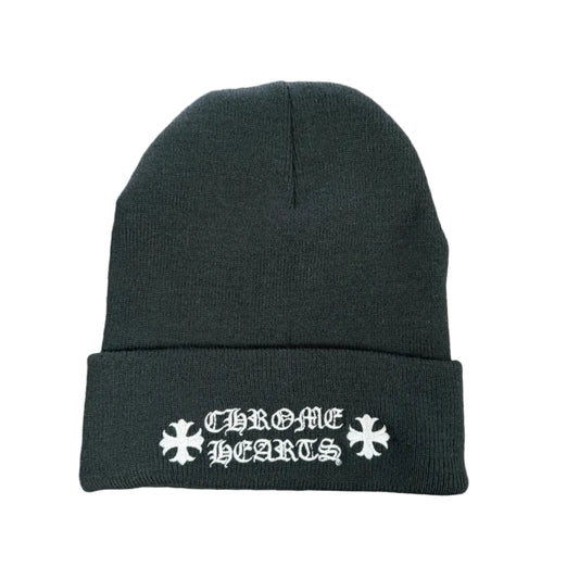 A black knit beanie with white embroidered gothic lettering and snowflake motifs on the cuff, laid flat on a round wooden surface.